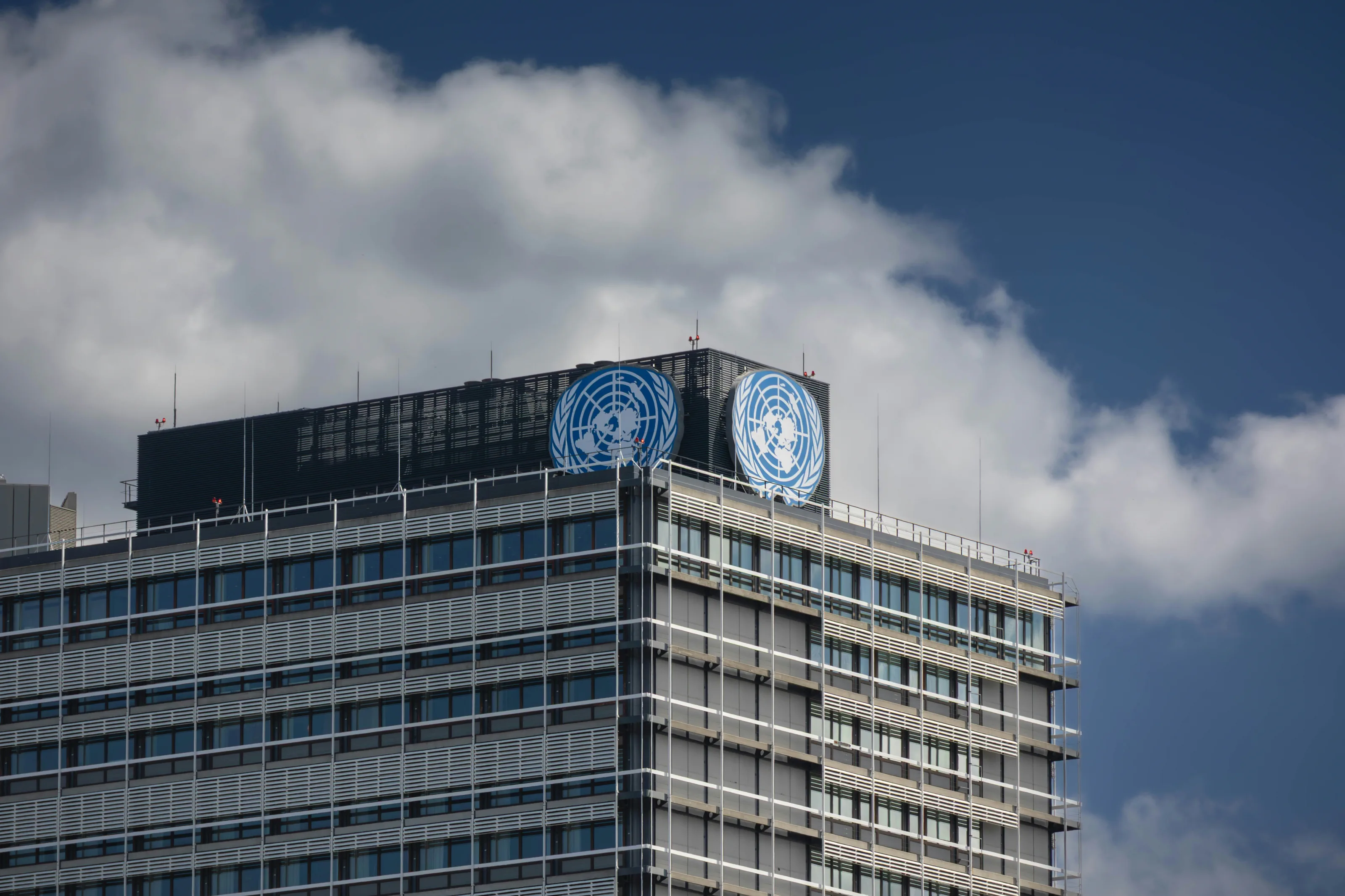 Gebäude der Vereinten Nationen mit zwei UN-Emblemen vor bewölktem Himmel.