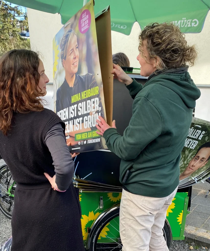 Zwei Personen befestigen ein Wahlplakat an einem grünen Lastenanhänger.